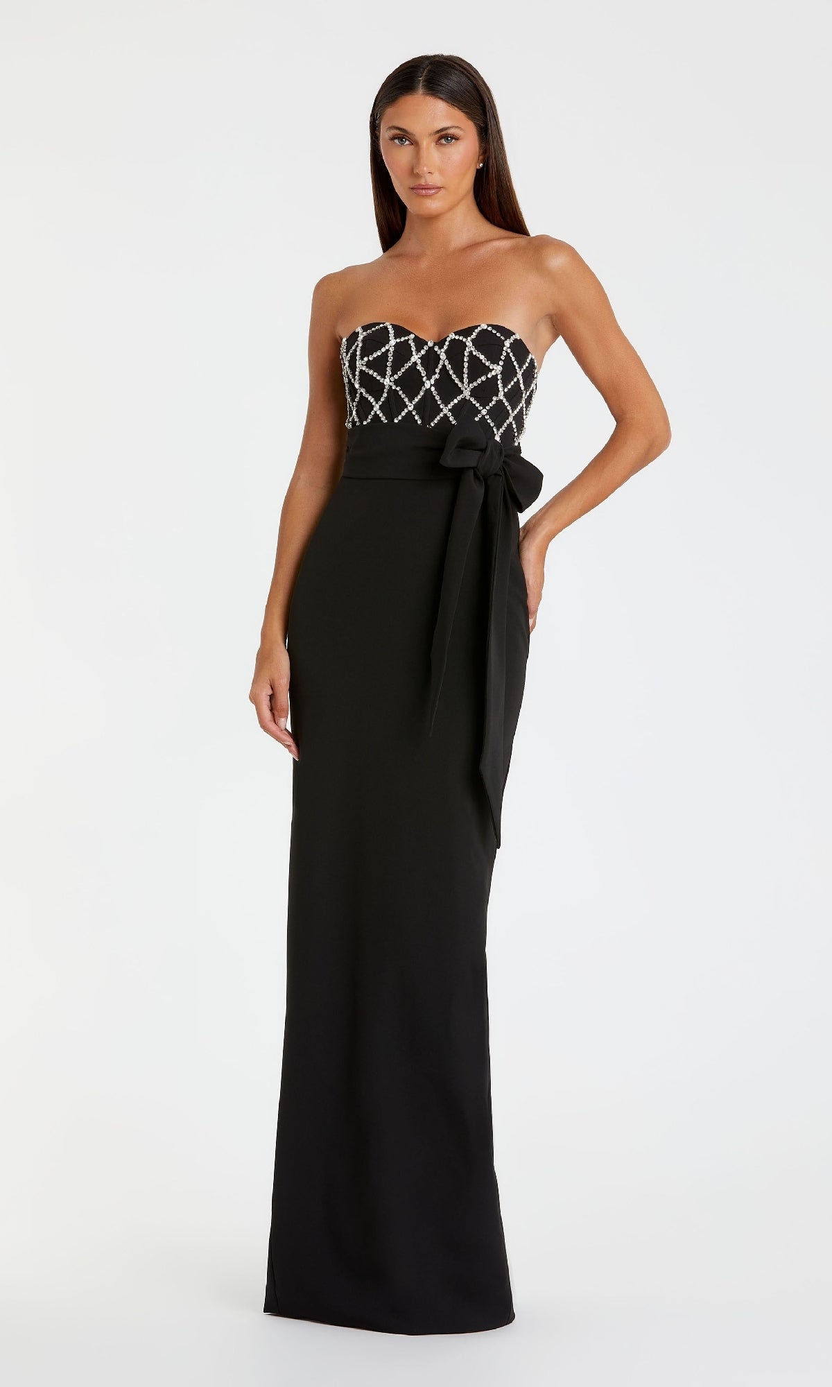 A woman models the Strapless Sweetheart Formal Gown: Mac Duggal 2294, featuring a geometric beaded bodice, waist bow, and flowing skirt, as she stands against a plain white background.